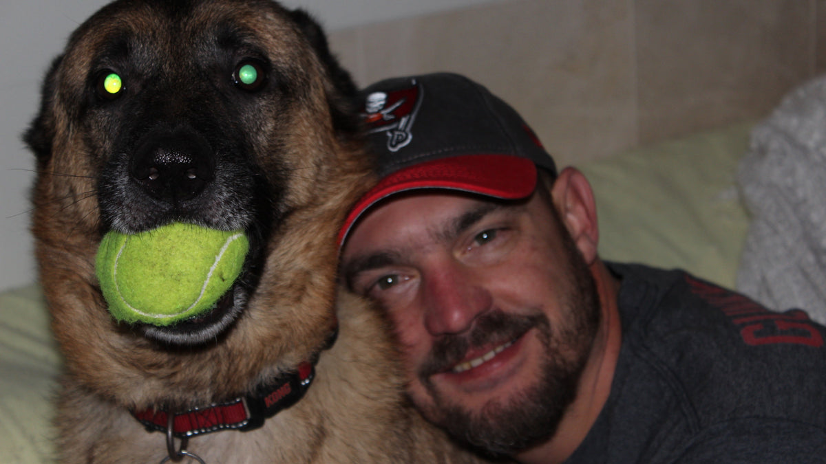 A large tan and black dog holding a bright yellow tennis ball in its mouth, posing closely with a smiling man wearing a Tampa Bay Buccaneers cap and dark gray shirt in a bedroom setting.
